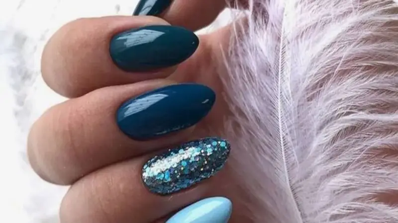 Unghie per l'inverno: unghie blu petrolio e azzurre con glitter scintillanti, accanto a una soffice piuma bianca.